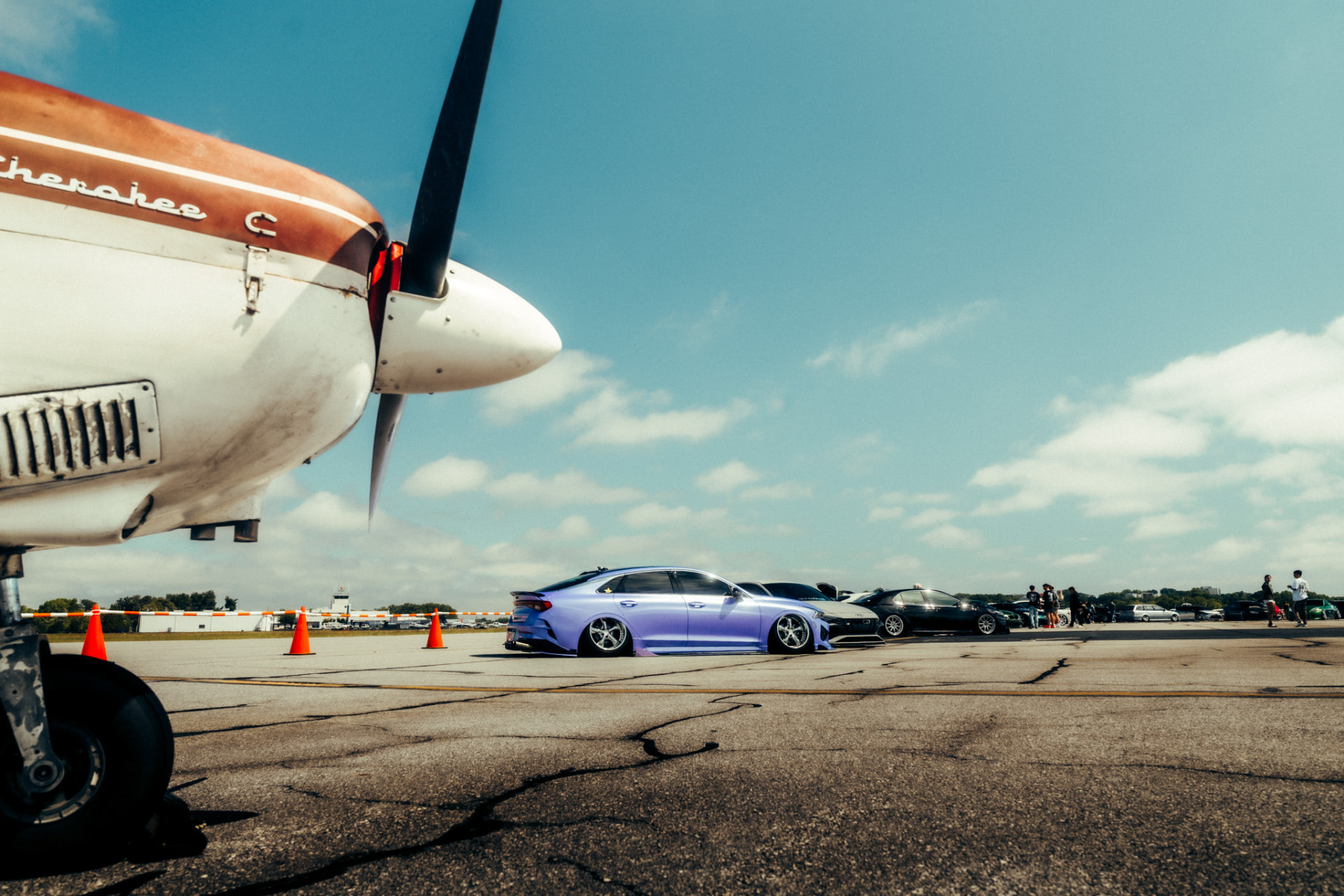 Lavender Kia K5 parked beside a Piper Cherokee propeller at an airport car show