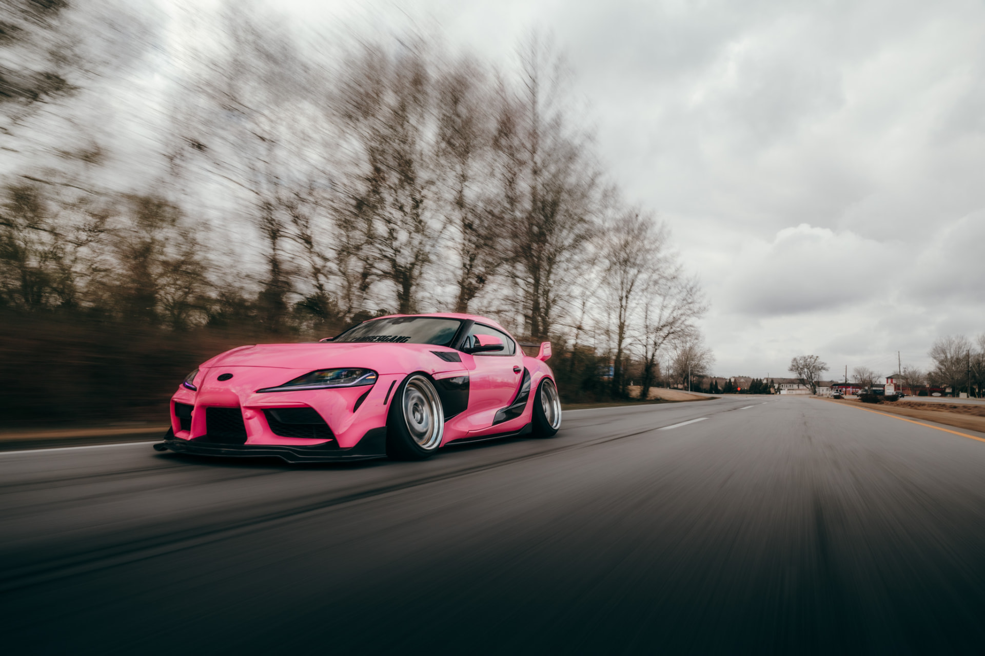 Pink Toyota Supra rolling down the highway with blurred trees behind