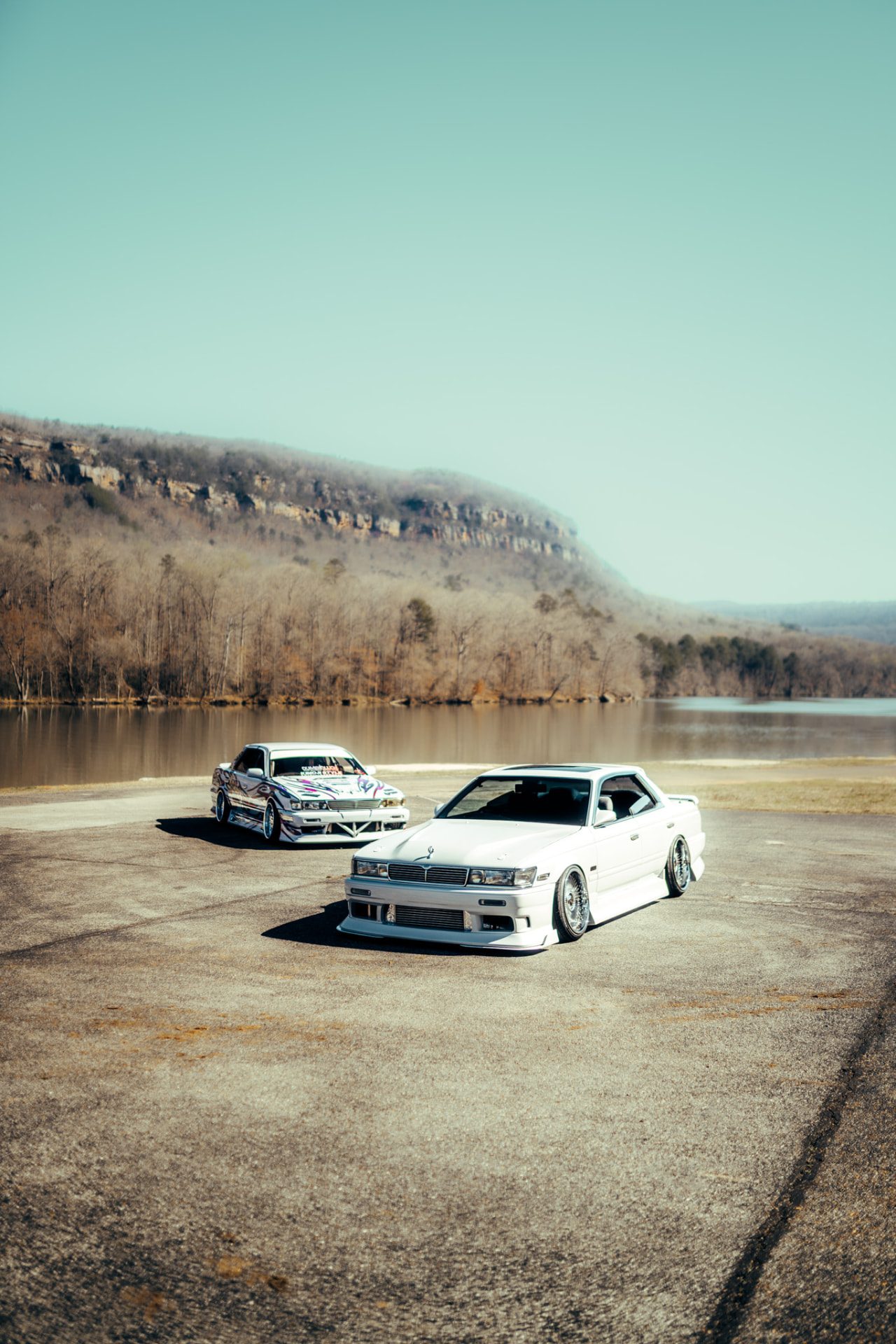 Two white JDM sedans parked on a lakeside lot beneath a mountain